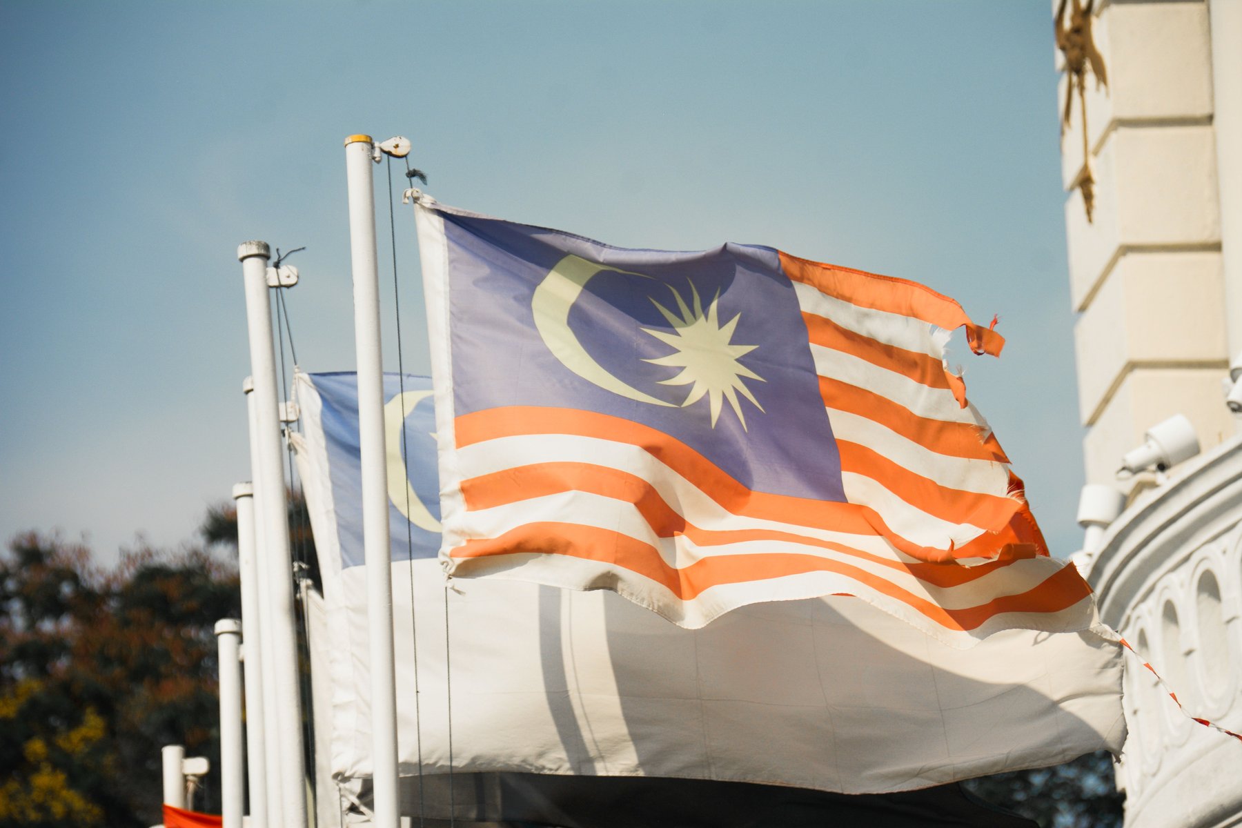 A sun-bleached Malaysian flag, frayed along its fly edge, flying from a white pole against a clear sky, with a second, paler flag on the pole behind it and the pale stonework of a colonial-era public building to the right